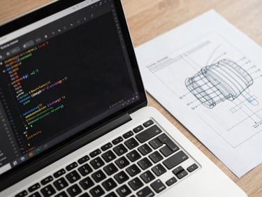 Overhead close-up of a desk with an open code editor on a laptop screen showing Kotlin Android code, a printed wireframe sketch beside it, natural daylight from above, no hands or faces, sharp technical detail