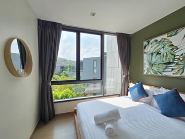 Modern hotel bedroom with white linens, blue pillows, and large windows overlooking lush greenery.