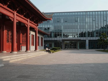 Wide shot of a modern Chinese university campus courtyard in morning daylight, traditional red-pillared architecture beside a contemporary glass library building, a single student reading on steps in the middle distance, natural light, documentary style