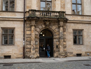 Wide shot of a historic Prague university building exterior on an overcast day, cobblestone street in the foreground, arched stone doorway, a student with a backpack entering the building, documentary natural light, no tourist staging