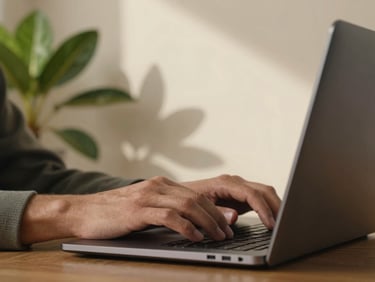 Close-up of a person's hands typing on a sleek laptop next to a green plant, warm natural Texas light casting soft shadows on a cream-colored wall.