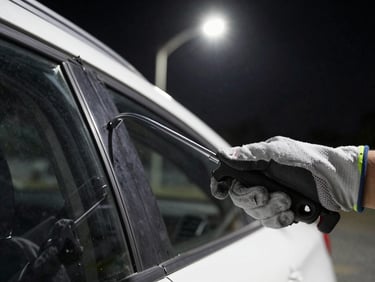 Tight shot of gloved hands using a slim-jim tool along a car door frame at night, streetlight overhead, tools visible in foreground