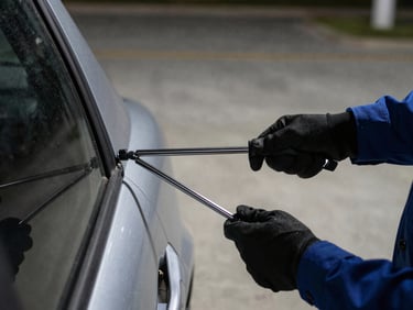 Technician's gloved hands working a slim-jim tool into a car door frame at night under a streetlight, vehicle door edge visible, concrete lot in background