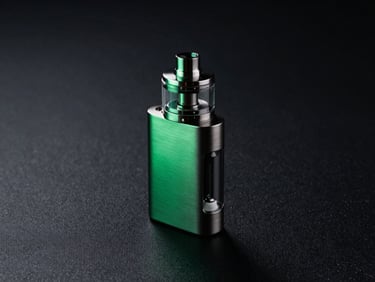 Premium brushed-metal vape kit with a clear glass tank, resting on a dark textured surface, dramatic emerald green backlighting
