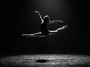 Wide frame, stage lighting from above, dancer mid-leap against deep black backdrop, full body in sharp silhouette, arms extended, motion blur on fingertips, high contrast monochrome with single amber gel catch light