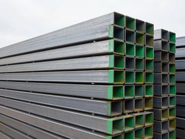 Stack of neatly organized, heavy iron beams in an industrial yard, clean metallic tones, bright overcast daylight, sharp focus, 35mm, corporate-industrial style
