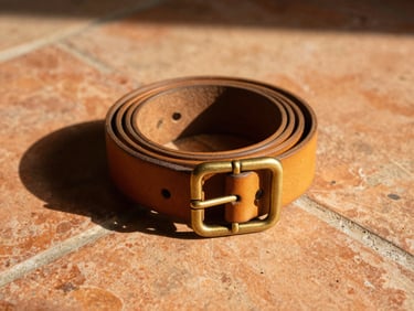 A rolled tan leather belt next to hand-cast brass buckles on a terracotta tile, warm golden lighting, sharp shadows.