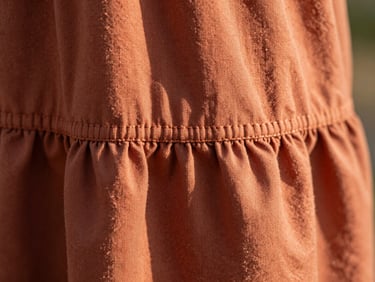 Detailed macro shot of a terracotta-colored cotton frock hem, showing neat hand-finished stitching, warm natural sunlight highlighting fabric texture, shallow depth of field.