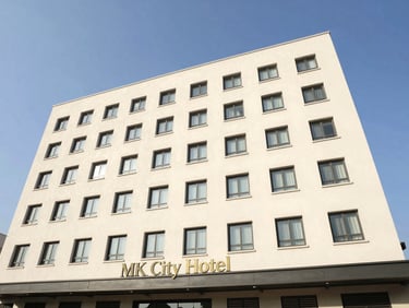 Bright daytime exterior photo of MK City Hotel, showing a clean multi-story building facade under a clear blue sky, realistic.