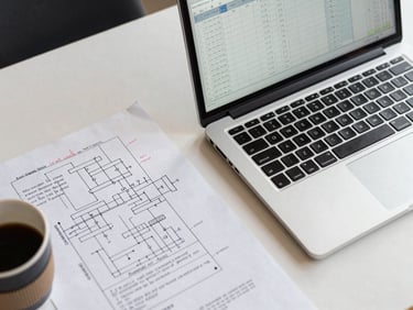 Close-up overhead desk study in office fluorescent light, a laptop open to a spreadsheet of operational data beside a printed process diagram with handwritten annotations, a coffee cup partially in frame, organized workspace, no people visible, clean and methodical atmosphere