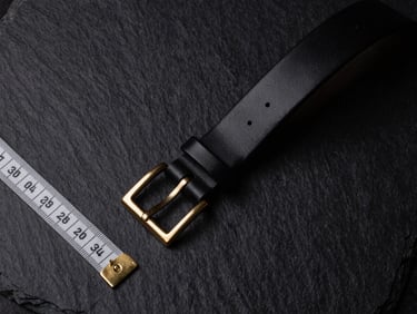 Overhead flat-lay of a measuring tape beside a matte black leather belt, with gold-tipped ends, on a dark velvet surface, low-key lighting.