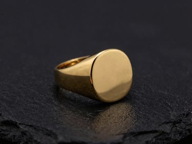 Tight macro shot of a heavy solid gold signet ring, sharp reflective gold highlights, emerging from deep shadows on a a matte black velvet surface, moody studio lighting.