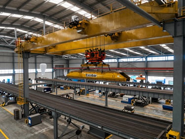 Wide drone shot of a modern steel fabrication plant in Escobar, showing heavy overhead cranes lifting massive steel trusses under natural daylight.
