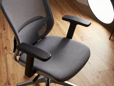 Close-up overhead shot of a high-back ergonomic mesh office chair on a hardwood floor, studio strobe lighting from the upper right revealing lumbar support structure and armrest adjustment detail; no person, clean neutral background