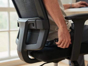 Close-up of a person's hand adjusting the lumbar support knob of an ergonomic office chair, bright daylight from a window to the left, focus on the hand and mechanism detail, neutral background