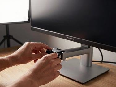 Detail shot of a monitor arm clamp attached to a desk edge, holding a widescreen monitor at eye level; hands adjusting the tilt angle, studio strobe from the upper left highlighting the aluminum finish and cable management channel