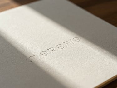A close-up macro shot of heavy cotton paper stationery with blind debossed typography, casting soft long shadows in warm morning window daylight.