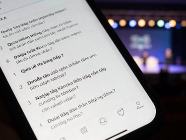 Overhead phone shot showing a Q&A submission interface with Vietnamese text questions listed, event stage blurred in background, ambient stage lighting