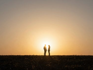 Silhouette of two figures standing close at the edge of a field, sun low and blazing behind them, sky wide and amber, figures barely distinguishable from the light surrounding them