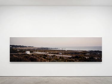 Wide panoramic photograph of a large-format archival pigment print from a coastal area with subtle human infrastructure, mounted on a clean white gallery wall, soft diffused daylight.