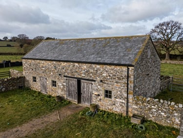 Overhead wide shot of a stone barn exterior in a rural UK setting, overcast even daylight, weathered timber doors, surrounding farmyard visible