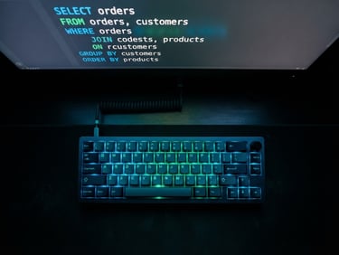Overhead shot of a mechanical keyboard glowing in a dark room, with a screen showing high-contrast SQL code syntax in neon blue and emerald green.