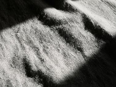 Extreme close-up of textured linen fabric under sharp, high-contrast sunlight with deep architectural shadows, monochrome desaturated tones, 35mm