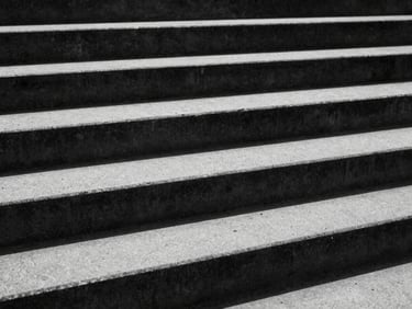 Extreme close-up of raw concrete architectural stairs under harsh, high-contrast sunlight with deep black shadows, 35mm, monochrome desaturated tones.