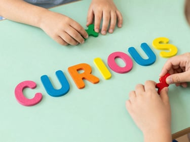 Vibrant wooden alphabet letters arranged on a mint green table spelling 'CURIOUS', with a child's hands visible in the frame, bright high-key editorial photography, cheerful natural daylight, vibrant primary colors, shallow depth of field