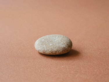 A single, smooth, light-colored stone resting on a terracotta-colored textile, soft diffused daylight, warm earth tones, minimalist composition.