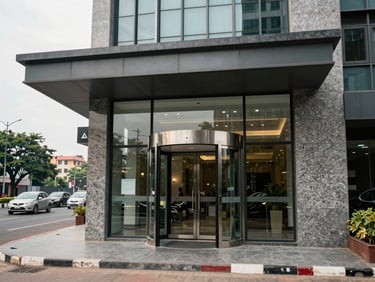 Street-level view of a modern commercial building entrance at Benz Circle Vijayawada, flat overcast daylight, glass revolving door, polished granite cladding, wide environmental shot showing the busy intersection context in the background