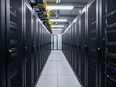 Wide shot of a data center corridor with two rows of server racks stretching into the distance, overhead fluorescent lighting illuminating the aisle, fiber optic cables bundled along the ceiling, no people, cool industrial environment, shot at mid-height looking down the aisle