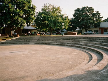 Warm editorial photography of an open-air amphitheater plaza, concrete steps, green trees around, late afternoon sun, 35mm film style.