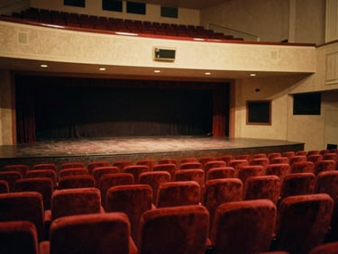 Warm editorial photography of an indoor theater stage with red velvet seats, dramatic stage lighting, 35mm film style.