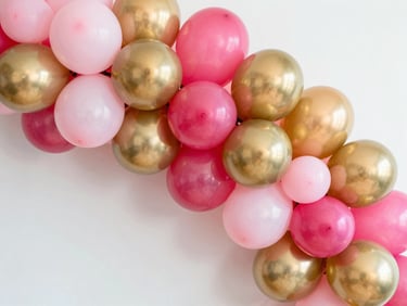 Close-up overhead of a lush pink and gold balloon garland arch, tightly clustered balloons in blush, hot pink, and metallic gold, bright studio lighting, white backdrop, sharp color saturation