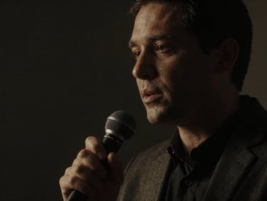 Dramatic low-key image of a male speaker with microphone, deep focus, chiaroscuro lighting, desaturated tones.