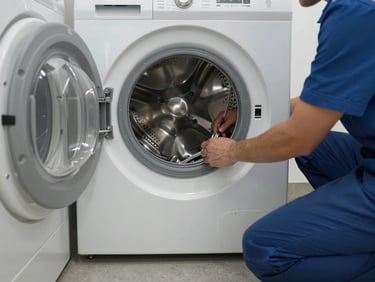 Technician kneeling beside a front-load washing machine, back panel removed, hands inspecting the drum belt under window light, concrete laundry room floor, no staging