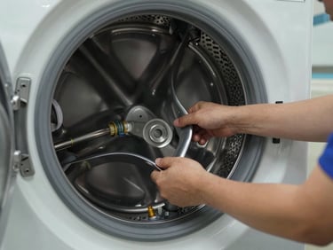 Technician's hands pulling a washing machine drum assembly in a laundry room, natural light from a doorway, worn drum seal and hose connections visible