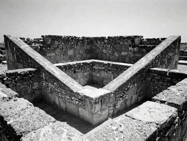 Stark documentary photography of a geometric stone structure built amidst ancient ruins, high-contrast monochrome, sharp midday shadows, 35mm film grain