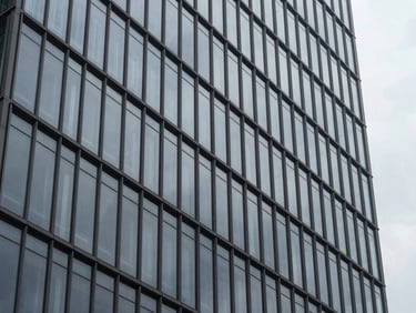 Close-up of a modern high-rise glazed facade reflecting an overcast sky, sharp architectural lines, desaturated steel and charcoal tones.