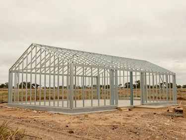 Architectural wide-angle shot of a 40x60 barndominium steel frame under construction, exposed metal trusses, concrete slab foundation visible, rural landscape under overcast daylight, 35mm lens