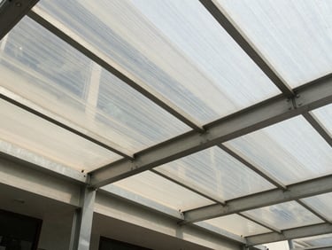 Wide environmental shot of a polycarbonate canopy structure over a building entrance in Indonesia, daylight filtering through the translucent cover, steel anchor bolts and main beam visible at the connection point