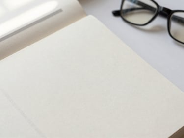 Overhead flatlay of an open minimalist book with clean typography on warm paper, soft window light, reading glasses nearby