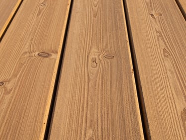 Tight environmental shot of hardwood decking boards laid out in natural afternoon light, grain lines running left to right, knot detail and colour variation visible across three adjacent boards, no furniture or people