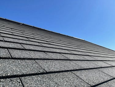 Low-angle dynamic shot of high-performance residential roof shingles catching bright direct afternoon sunlight, showing sharp clean lines against a vivid blue sky.