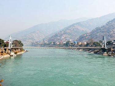 Panoramic view of Rishikesh suspension bridge over the turquoise Ganges river, misty mountains in the background, bright daylight.