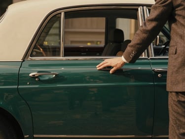 Intimate detail of a private chauffeur opening the door of a vintage luxury car, soft natural sunlight, rich emerald and beige tones.