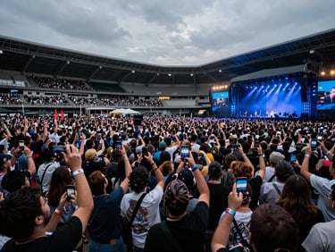 Wide overhead shot of a packed outdoor event arena, 5000+ attendees with smartphones and wristbands visible, cool blue stage lighting casting long shadows, desaturated industrial sky, no celebratory framing — operational scale