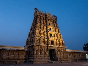 Wide shot of a Tamil Nadu temple gopuram at dusk, dramatic layered stone carvings catching last light against a deep blue sky, empty forecourt in the foreground, no tourists