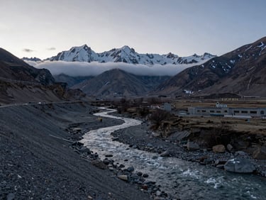 Wide environmental shot of Spiti Valley at early morning, high-altitude river cutting through grey scree, snow-dusted peaks in the far distance, low horizontal cloud layer, no people